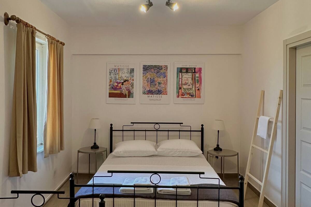 A bedroom with a bed, two bedside tables, a window with curtains, and three wall-mounted artworks.