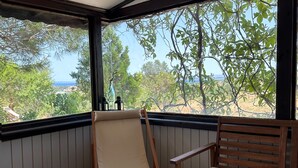 A covered patio area with a wooden bench and a cushioned seat, a table with a magazine and a vase of flowers, and a view of greenery and a distant landscape. - Denizhan Bağ Evi (Bozcaada)