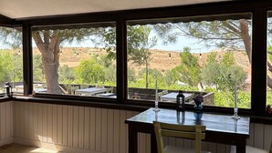 A room with a wooden table and chairs, large windows with a view of a tree and landscape. - Denizhan Bağ Evi (Bozcaada)