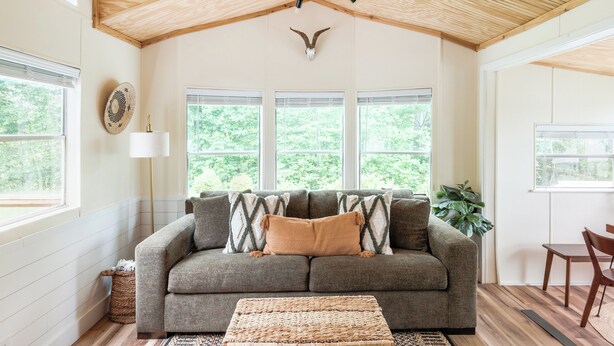 Una sala de estar acogedora con techo de madera, un sofá gris con cojines estampados, una mesa de centro trenzada y un ventanal con vistas a zonas verdes.