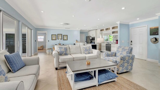 A spacious living room with a sofa, armchair, and coffee table, a kitchen in the background, and a sliding glass door leading to a balcony.