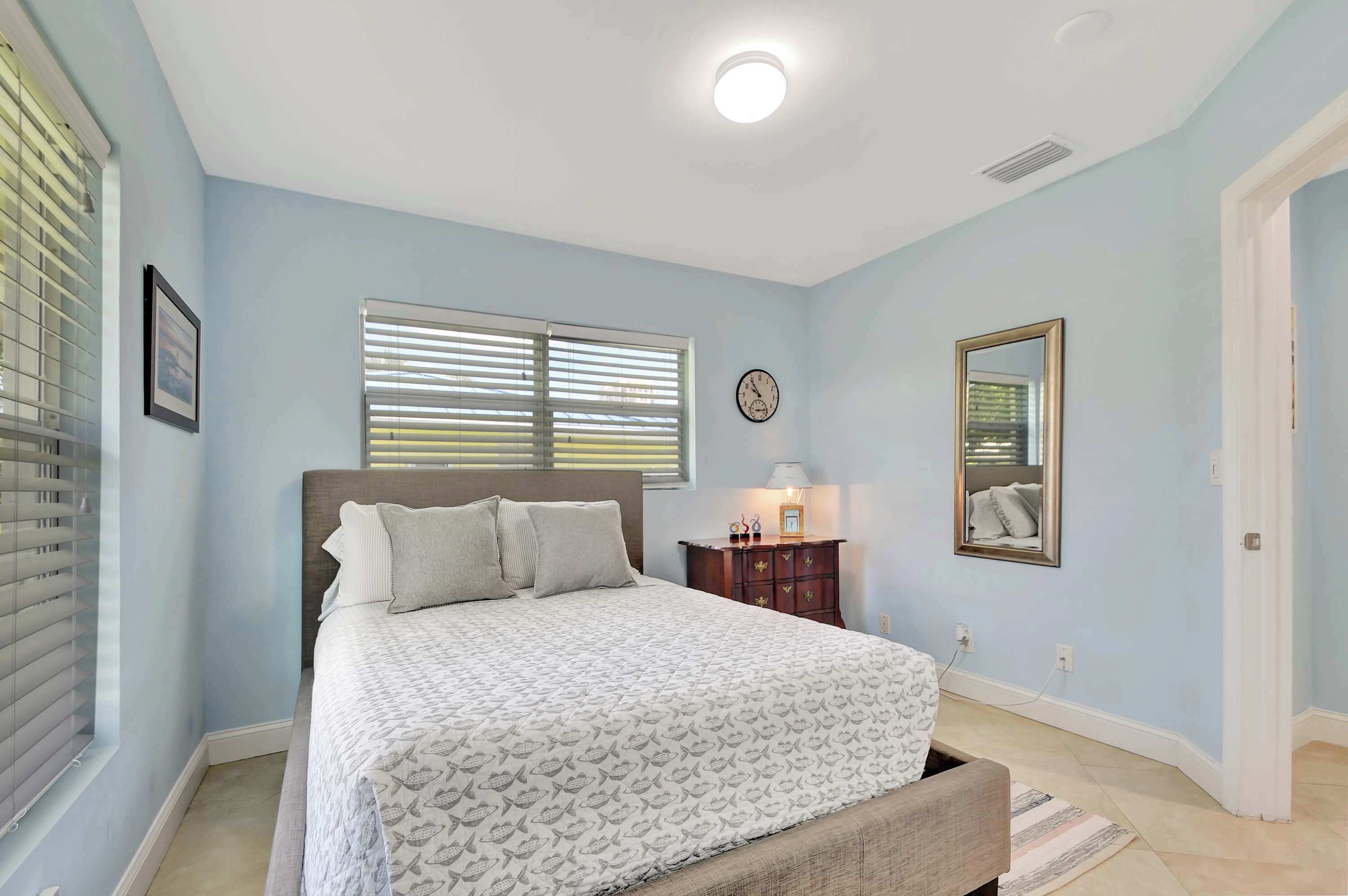 Une chambre à coucher comprenant un lit, une table de chevet, une commode, une horloge, un miroir et une fenêtre avec des stores.