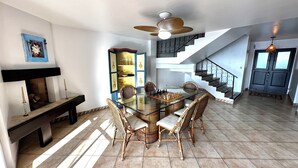 Uma sala de jantar com mesa de vidro cercada por cadeiras de vime, lareira e escada que dá acesso a um andar superior.