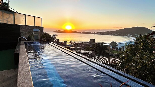 Piscina com vista para o pôr do sol sobre o mar, uma parede de pedra e um edifício moderno com grandes janelas.