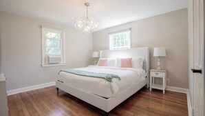 A bedroom with a white bed, two bedside tables, a chandelier, and two windows. - Family Cottage with 4 King Beds and Fire Pit, Norwalk/Westport Border (Norwalk)