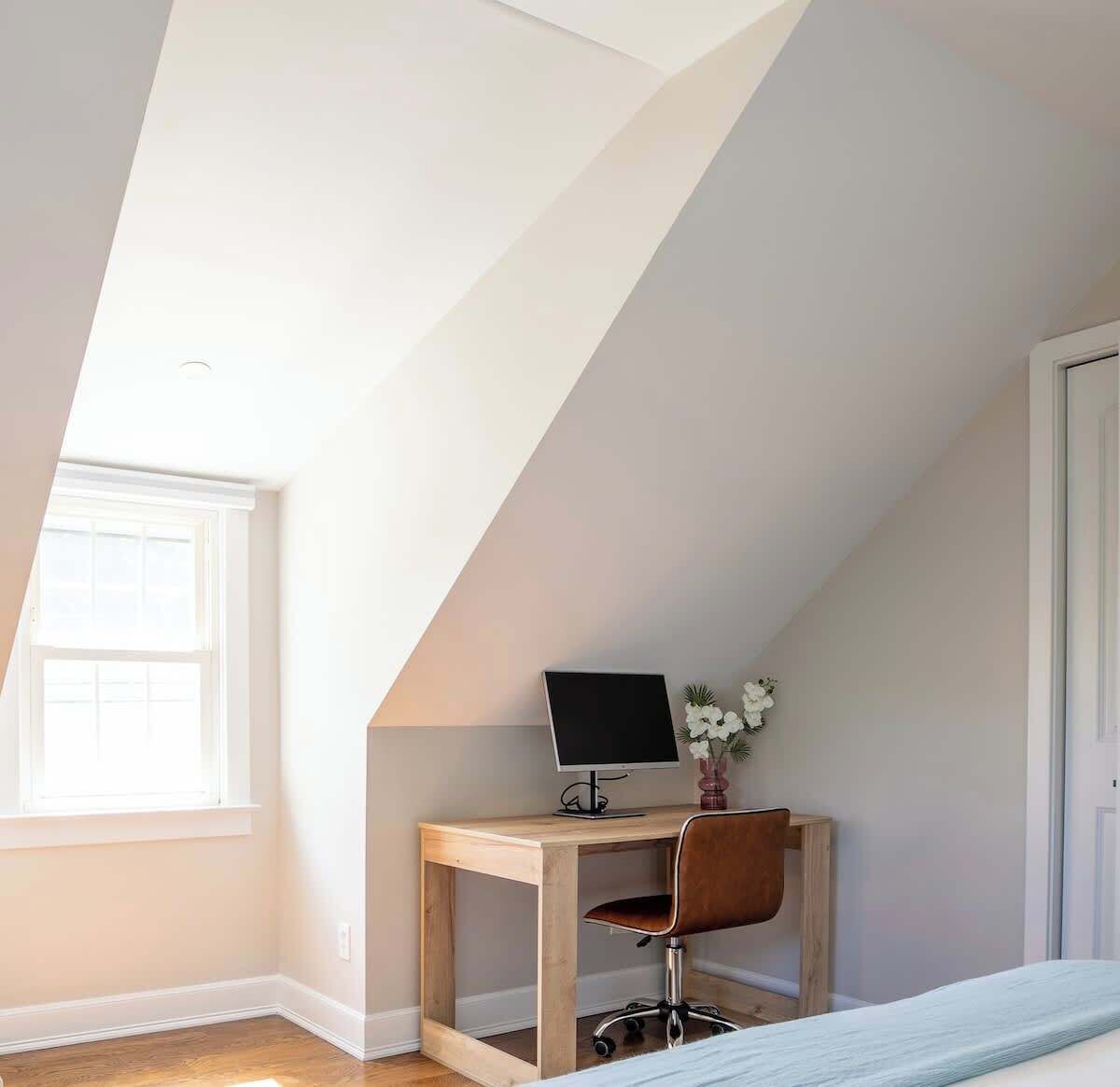Een slaapkamer met een schuine plafond, een houten bureau met een computer, een stoel en een bed met een blauwe deken.