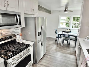 A kitchen with a microwave, stove, refrigerator, and dining area with chairs and a table. - Greenbelt Guesthouse (Long Beach)