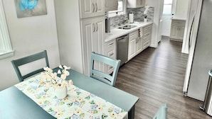 A kitchen with white cabinets, a stainless steel refrigerator, and a dining area with a table set for two. - Greenbelt Guesthouse (Long Beach)
