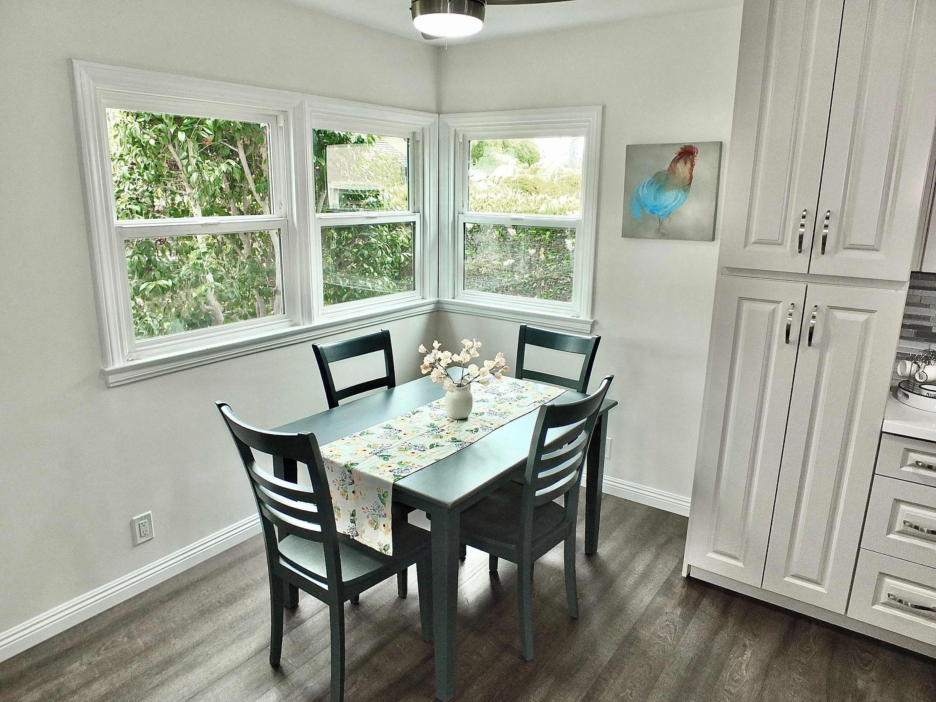 A dining area with a table set for four, surrounded by dark wooden chairs. There are three windows providing natural light, and a painting of a bird hangs on the wall.