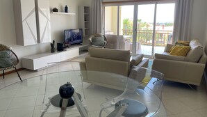 En moderne stue med et sofabord i glas, en sofa, et fjernsyn og en balkon med udsigt.