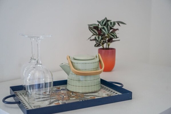 Una copa de cristal transparente con vino, una tetera verde con asa de madera y una planta en maceta roja sobre una bandeja.