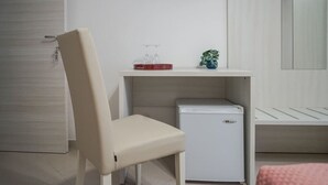 Une petite pièce propre, avec un bureau blanc, une chaise, un petit réfrigérateur blanc et un téléviseur fixé au mur.