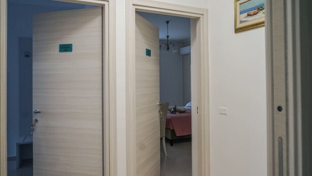 Un couloir avec deux portes : l’une est marquée de l’inscription « CHAMBRE DE NUIT », et à travers l’ouverture de l’autre, on aperçoit un lit et une couverture rouge.