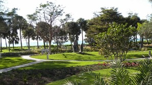 A landscaped garden with a winding path, various trees, and a view of the sea. - Beautiful Naples home, walk to private beach! (Naples)