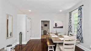 Un espace repas avec une longue table, des chaises blanches et un banc. Il y a un miroir au mur, un tableau représentant un taureau et une fenêtre avec des rideaux à motifs.