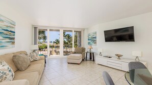 A spacious living room with a sofa, armchair, and a glass table, a flat-screen TV mounted on the wall, and a balcony with a view of palm trees and a pool. - Escape to paradise at our beachfront condo at the Gulfview. (Marco Island)