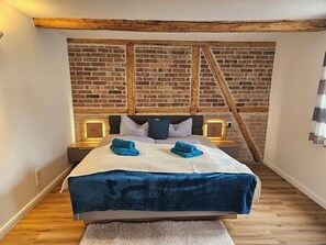 A hotel room with a brick wall, wooden beams, a bed with a blue blanket, and bedside lamps. - Urlaub im Harz - Ferienwohnung im Ilsetal mit Großer Terrasse (Ilsenburg (Harz))