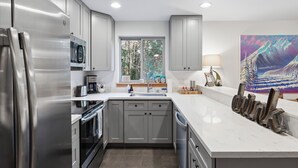 Cocina moderna con gabinetes grises, electrodomésticos de acero inoxidable, un amplio ventanal y un cuadro de un paisaje montañoso.