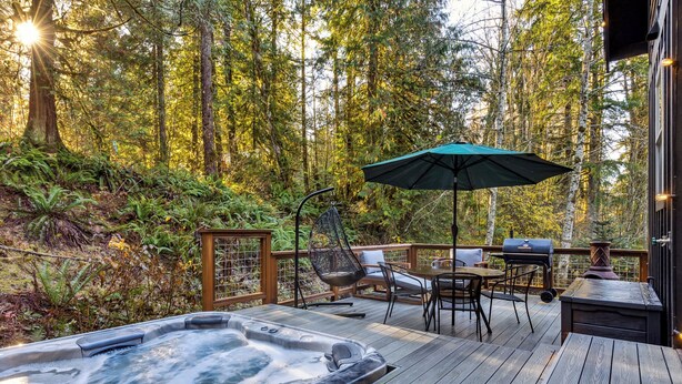 Un jacuzzi en una terraza de madera rodeado por un bosque, con mobiliario exterior y una parrilla.