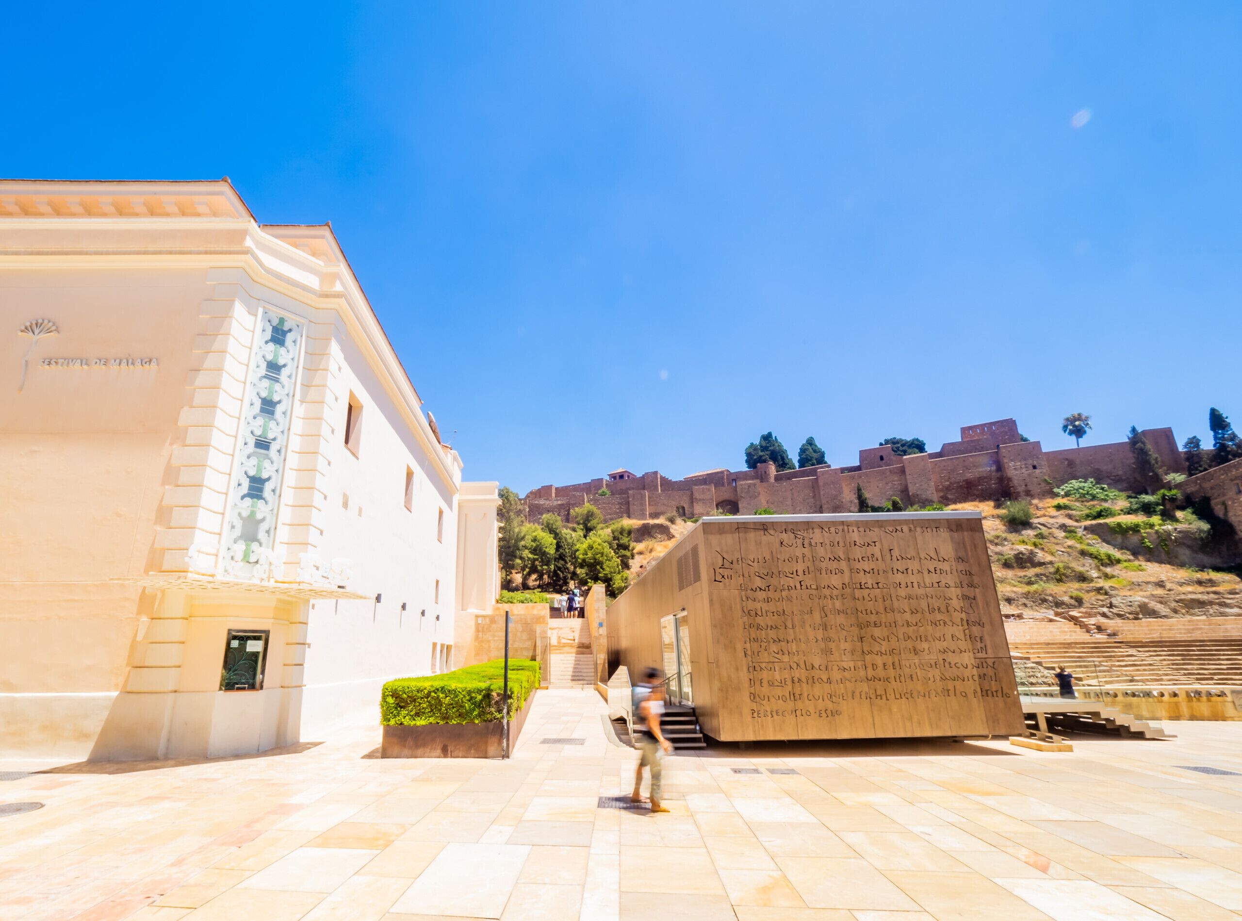 Un edificio histórico con cielo despejado, un gran monumento de piedra con inscripciones y un camino empedrado que lleva a una fortificación.