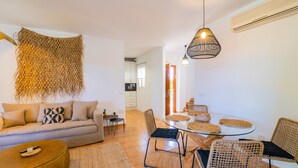 A modern living room with a sofa, coffee table, and wicker chairs. There is a dining table with placemats and chairs. A woven wall hanging and a hanging light fixture are visible. - Cubo's Townhouse Sierrezuela / communal pool / BBQ / terrace / parking (Mijas Costa)