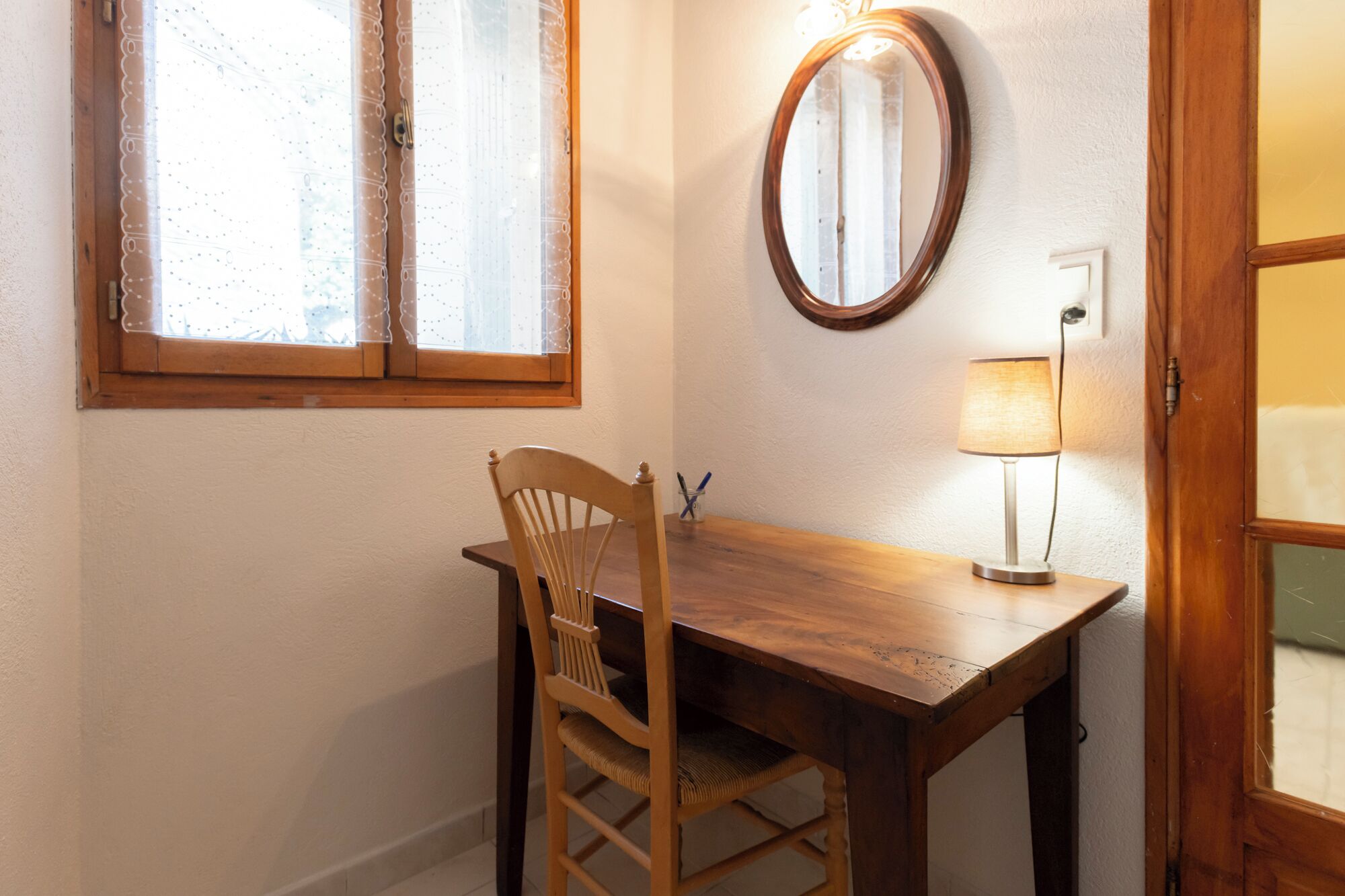 Une table à manger en bois, accompagnée d’une chaise, d’un miroir rond fixé au mur et d’une lampe de table.