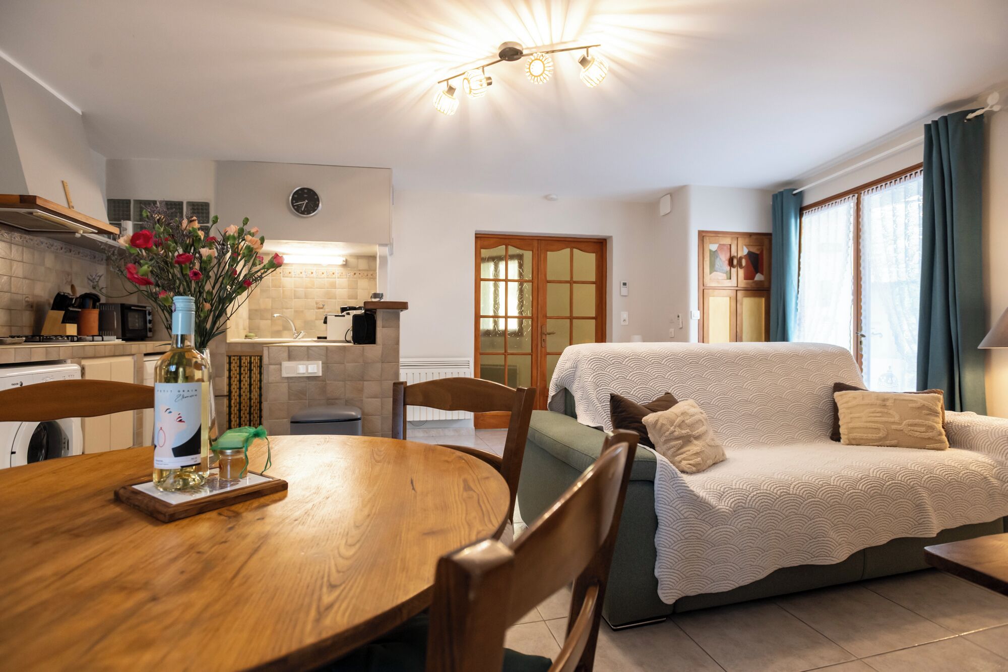 Un salon confortable avec un canapé, une table à manger et des chaises, ainsi qu’un coin cuisine agrémenté d’une horloge et d’un vase de fleurs.