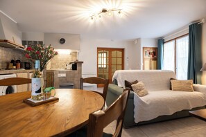 Un salon confortable avec un canapé, une table à manger et des chaises, ainsi qu’un coin cuisine agrémenté d’une horloge et d’un vase de fleurs.