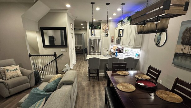 Una cocina moderna con armarios blancos, un comedor con mesa y sillas de madera, y una sala de estar con sofá gris y un banco.