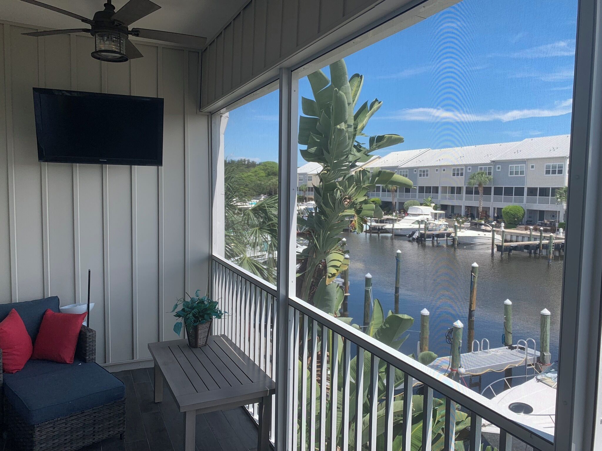 A balcony with a view of a marina, a television mounted on the wall, a wicker sofa with red cushions, and a small table with a plant.