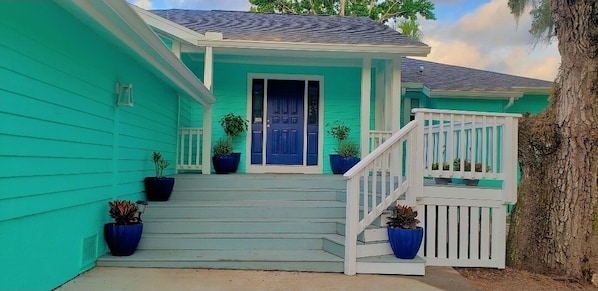 Uma casa turquesa com porta azul, corrimão de varanda branco e degraus até a porta da frente.