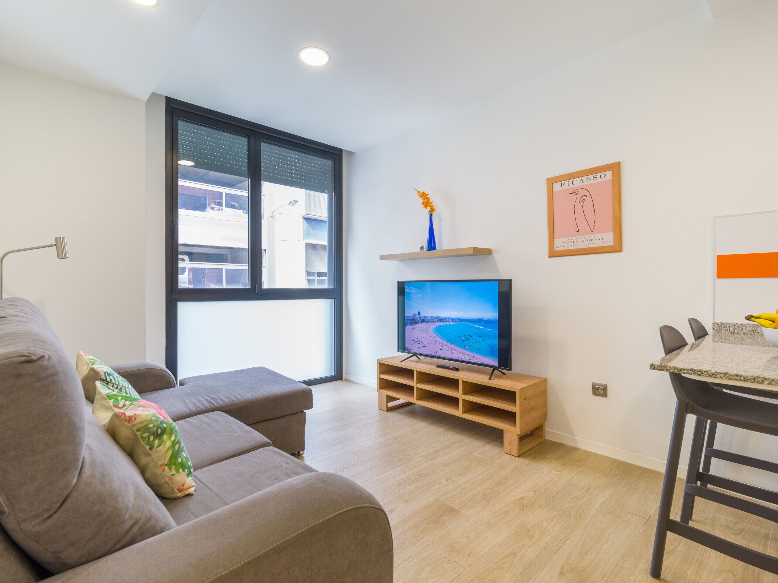 A modern living room with a grey sofa, a TV on a wooden stand, a dining area with a table and chairs, and a large window with a view of buildings.