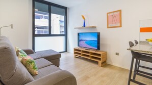 Ett modernt vardagsrum med en grå soffa, en tv på ett träställ, en matplats med ett bord och stolar samt ett stort fönster med utsikt över byggnader.