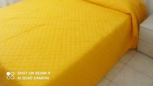Ein Schlafzimmer mit einem weißen Bett, einer gelb gesteppten Bettdecke und zwei grünen Wandbildern mit Blättern.