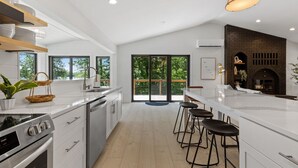 A modern kitchen with white cabinetry, a central island with bar stools, a brick fireplace, and large windows offering views of greenery. - Lake House w/Hot Tub, Game Room, Fireplace & Beach (Crosby)