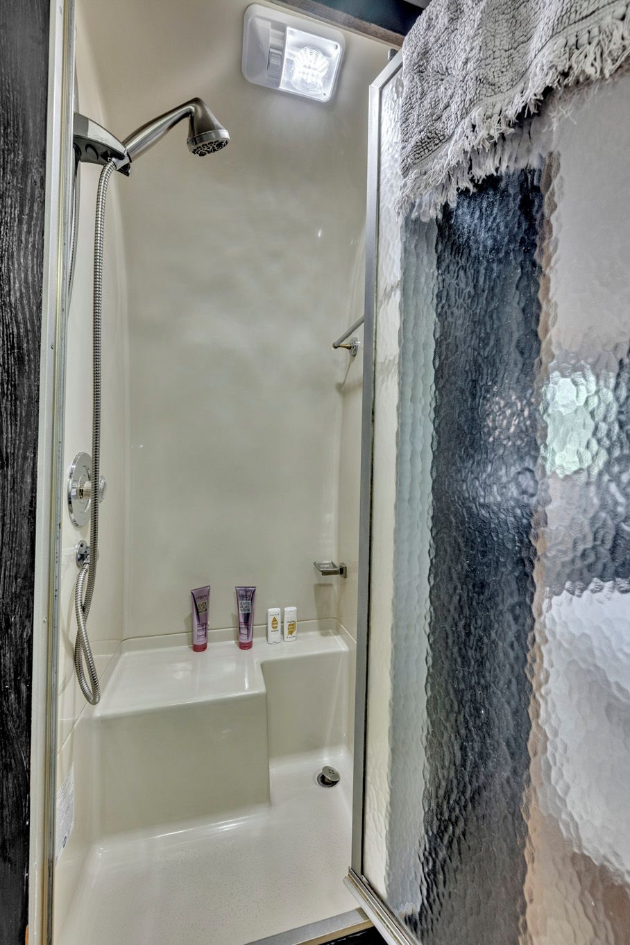 Une douche équipée d’un pommeau de douche à main, d’une paroi de douche en verre et d’une étagère pour les articles de toilette.