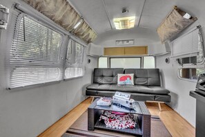 Interior de una caravana compacta, con un sofá, una mesita con revistas y una ventana con persianas.