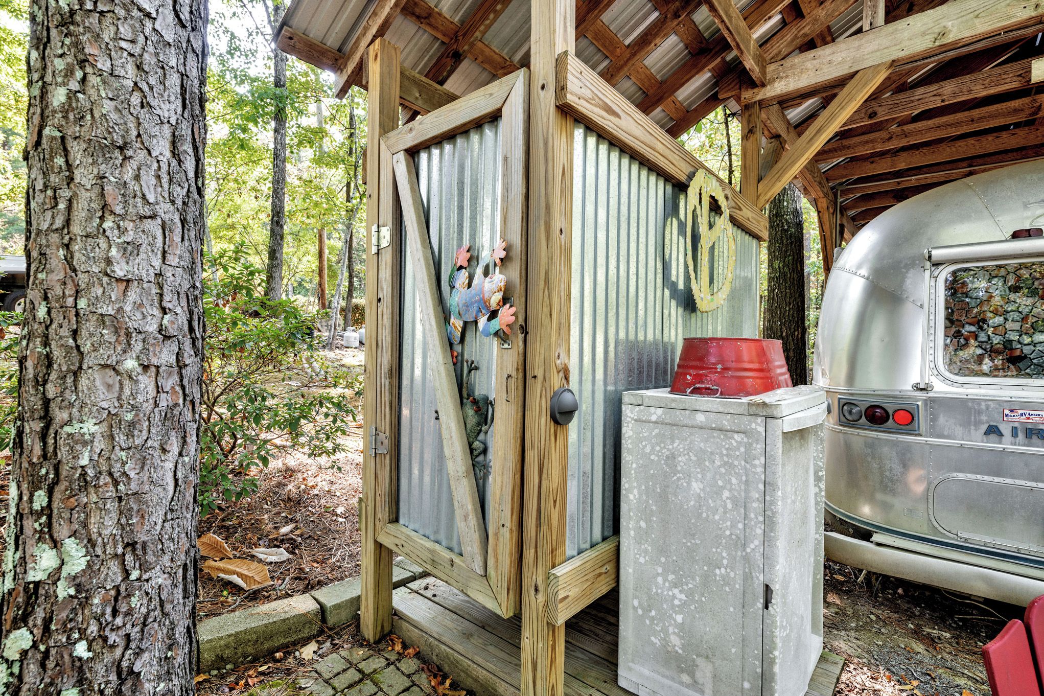 A rustic wooden structure with a corrugated metal door, a small concrete utility box, and a vintage RV parked nearby.