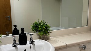 A bathroom with a white sink, two black soap dispensers, a small green plant, and a mirror reflecting a bathtub.