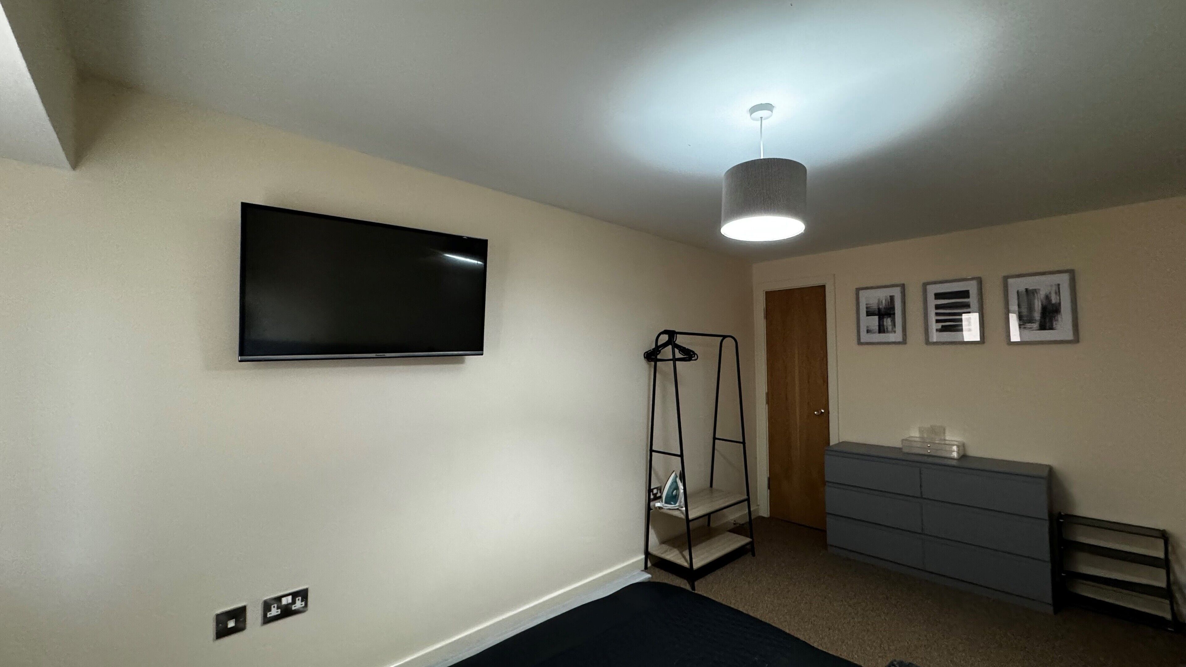 Een slaapkamer met een wandtelevisie, een kledingkast, een bed, een wasrek en een deur.