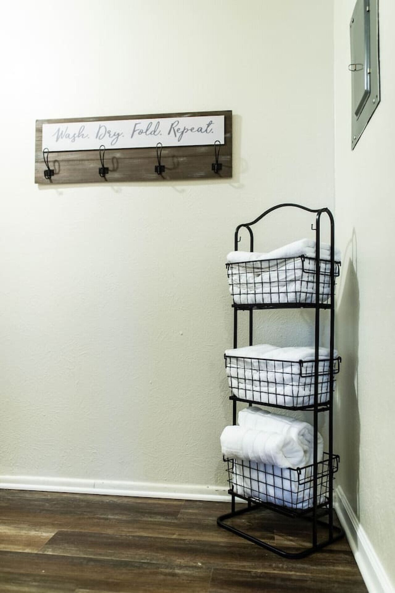 A metal wire shelf unit with three compartments, each holding rolled towels, against a white wall. Above it, a wooden sign with the text "Wash. Dry. Fold. Repeat." is mounted.