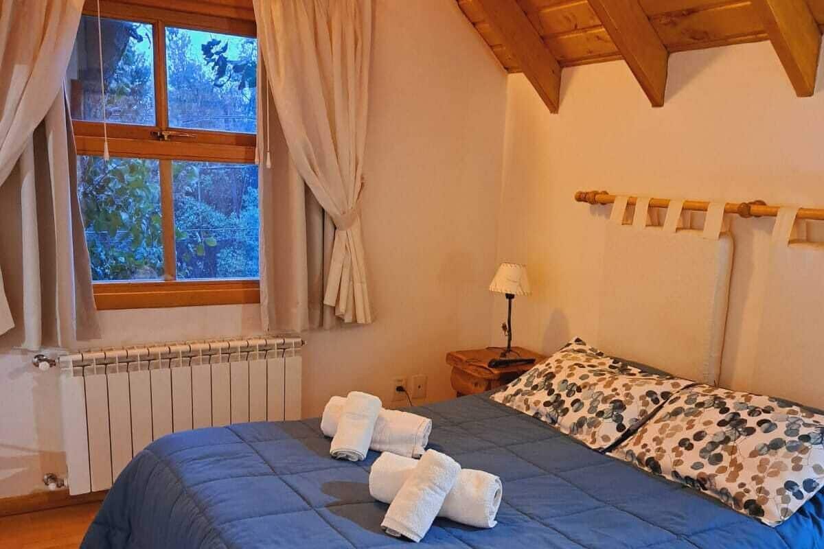 Quarto com teto de madeira, uma janela grande com cortinas, cama com colcha azul e toalhas brancas, e mesa de cabeceira com luminária.