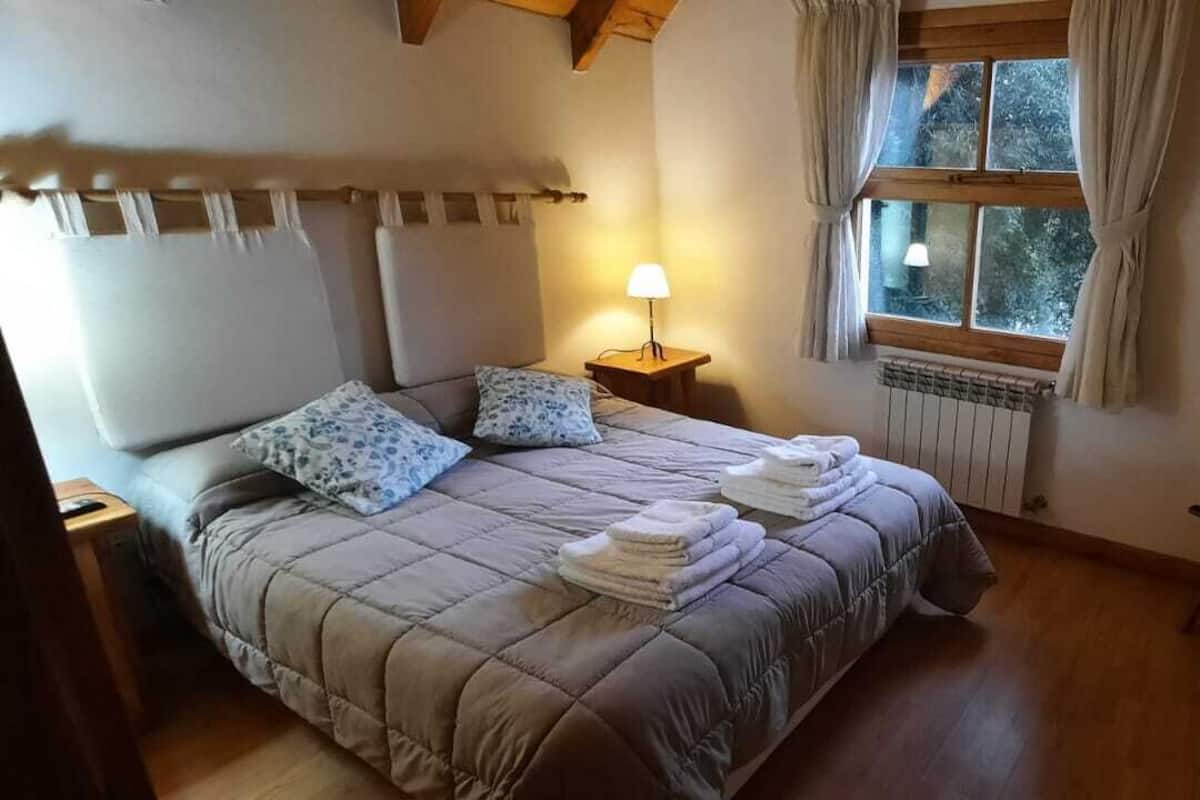 Quarto com cama, luminária de cabeceira, janela com cortinas, radiador e um banquinho de madeira.