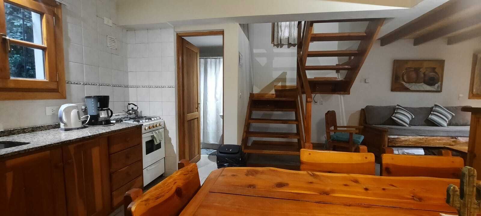 Cozinha com armários de madeira, fogão e máquina de café. Há uma mesa de jantar de madeira com cadeiras. Uma escada leva a um mezanino com vista para o exterior.