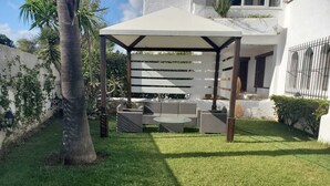 A covered outdoor seating area with wicker furniture and a table, surrounded by a well-maintained lawn and a white building in the background. - Apartment 2 min walk from cabo negro beach. (Tetouan)
