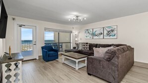 Een moderne woonkamer met een grijze bank, een blauwe fauteuil en een salontafel. Drie kunstwerken hangen in lijsten aan de muur, en er is een groot raam met uitzicht.
