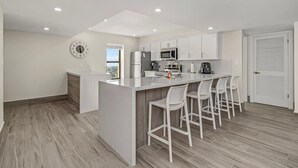 Een moderne keuken met witte kasten, een centraal eiland met barstoelen, een ingebouwde magnetron en een koelkast.