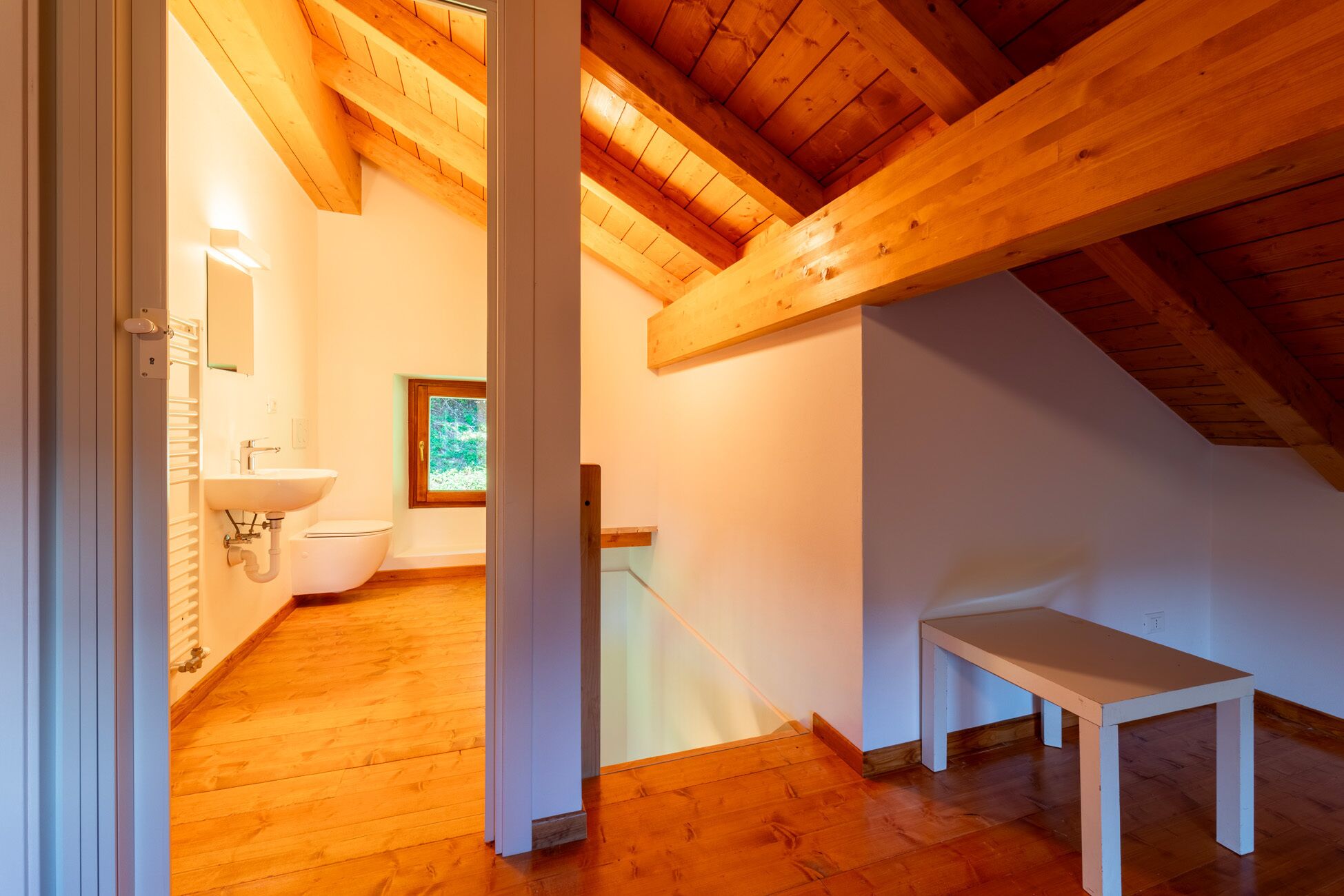 Una camera da letto in mansarda, con pavimento in legno, una finestra a lucernario, un lavandino bianco e una panchetta in legno.