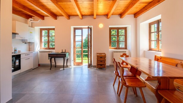 Una cocina amplia con vigas de madera, un comedor con una mesa grande y sillas, y ventanas grandes que ofrecen vistas a la vegetación.