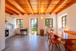 A spacious kitchen with wooden beams, a dining area with a large table and chairs, and large windows offering views of greenery.